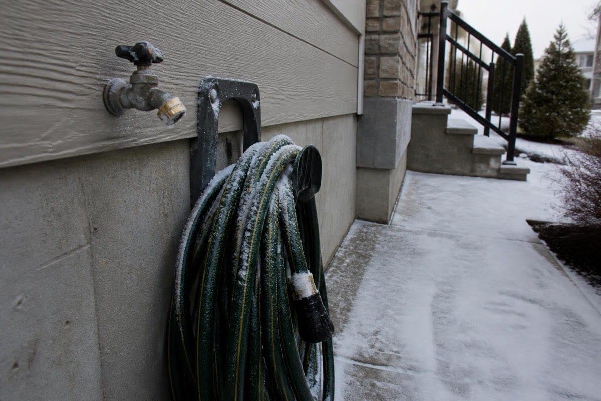 Snow-dusted suburban home exterior with a disconnected garden hose coiled beside an exterior hose bib
