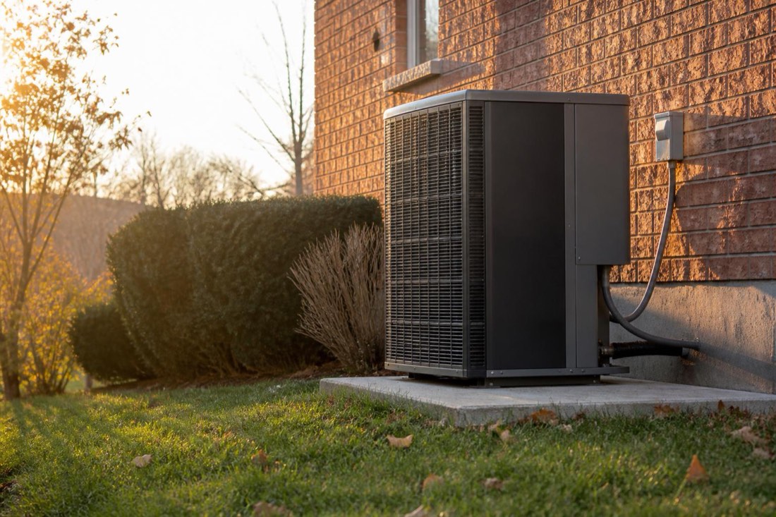 Outdoor heat pump condenser unit installed on a concrete pad beside the brick exterior wall of a suburban home at golden hour.