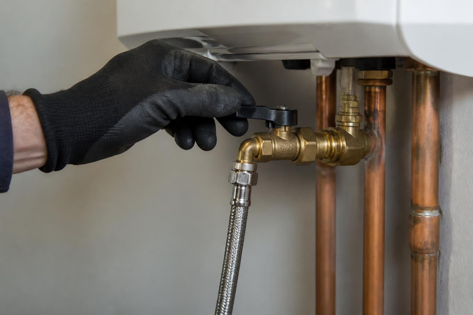 Gloved hand turning a shutoff valve on a wall-mounted boiler's copper pipe connections